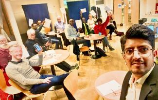 A man taking a selfie, with a group of people sitting at tables holding up papers and mobile phones