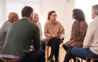 Adults in a support group setting, sitting in a circle. A woman is talking and others are listening to her