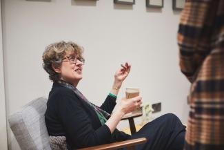 A woman in a chair chatting with somebody standing up, whose face we can't see