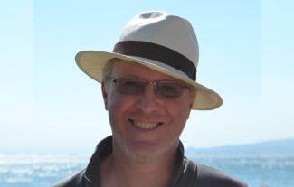 A man in a hat standing with the sea in the background, smiling for the camera