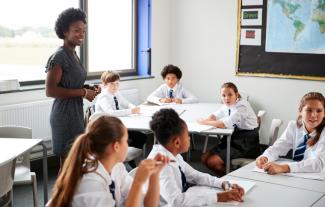 A teacher standing in a classroom, with pupils sitting on tables around her