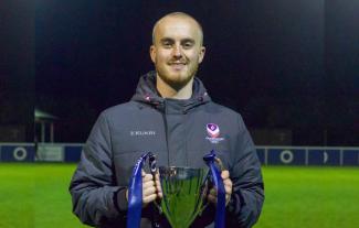 A man standing on a football pitch, holding a trophy and smiling