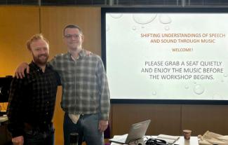 Two men standing beside a projection screen, with their arms around each other