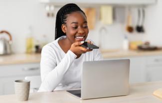 A woman sitting at a kitchen table in front of a laptop and speaking into a smartphone in her hand