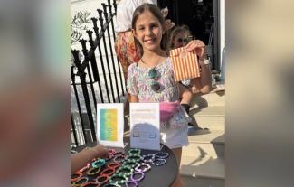 A girl smiling and holding up a paper bag, standing at a stall displaying bracelets