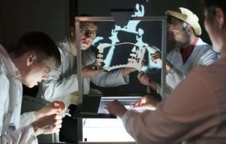 Four people in white lab coats sitting round a table, working on making puppets. Two are holding a puppet