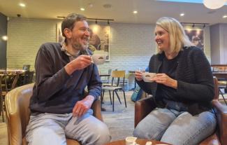 A man and woman sitting in a coffee shop and chatting, both holding cups of coffee