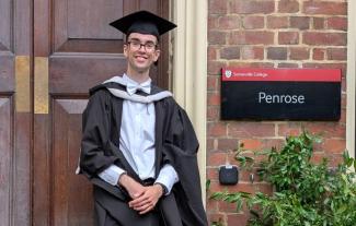 A young man in graduation robes, standing in front of a university door