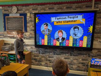 A boy standing at the front of a classroom next  to a digital screen, while other pupils watch on