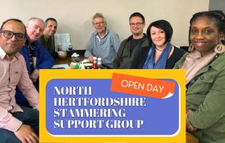 A group of people sitting in a cafe, smiling, with a sign saying 'North Hertfordshire Stammering Support Group Open Day' under them