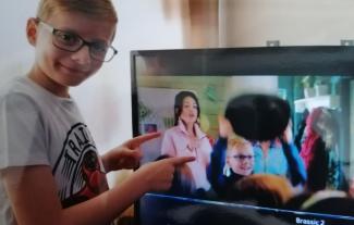 A boy smiling and pointing at a TV screen, on which he his being displayed.