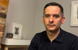 A man looking serious, in front of a living room mantlepiece