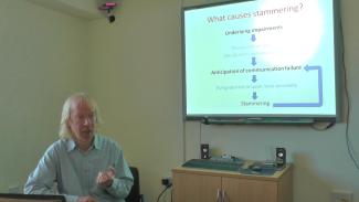 A man speaking at a desk in front of a screen with a diagram on it