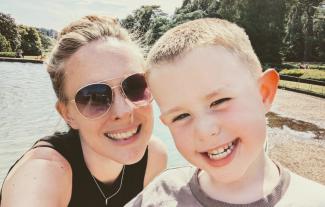 A woman wearing sunglasses standing next to her young son, in front of a lake. Both are smiling