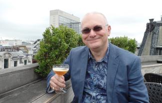 A man wearing sunglasses on a rooftop, smiling and holding a glass full of drink