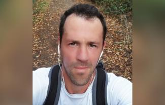 A man in a woodland setting, with earphones in and smiling