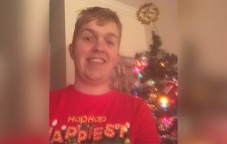 A man wearing a Christmas jumper, standing in front of a Christmas tree, smiling