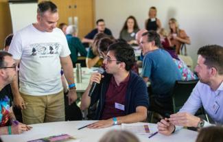 A man sitting at a table at a conference and talking into a microphone. Two men are sitting around him and a man is standing next to him, listening