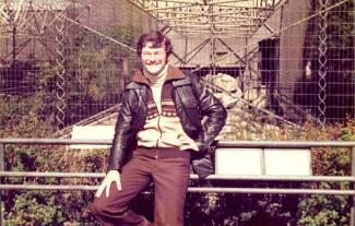 A man in 1970s clothing posing against a fence