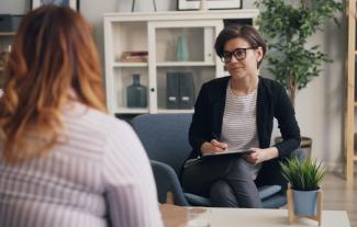 Two women in a counselling session, one sitting opposite the other making notes in a notebook.
