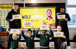 Two men standing either side of a screen, with four schoolchildren kneeling, all holding up paper with writing on it