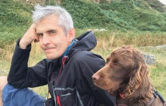 A man sitting on a hillside, resting his head on his hand, with his dog sitting next to him