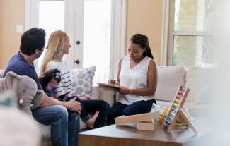 A father and mother sitting on a sofa with a young daughter between them, siting opposite a therapist who is taking notes