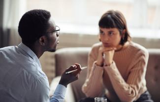 A man talking, with a woman listening to him, looking at him intently with her head resting on both hands