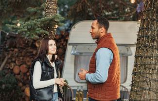 A man and a woman in an outdoor setting, standing and facing each other. The man is talking as the woman is listening.