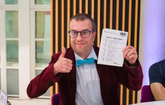 A man wearing a suit and a bow tie holding up a piece of paper and giving a thumbs up