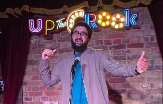 A man on a stage, smiling with his arms outstretched, in front of a microphone. The words 'Up the crook' are in lights behind him