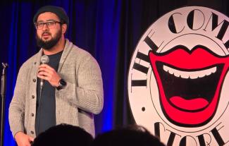 A man on stage speaking into a microphone. Behind him is a sign saying 'Comedy Store'