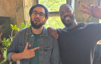 Two men smiling, one pointing to the other and the other man making a peace sign with his hand