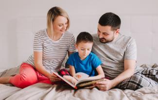 A mother and father sitting on a bed, with their young son between them, reading a book