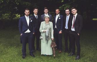 A woman standing in an outdoor setting, with six men standing behind her, all smiling and in wedding outfits