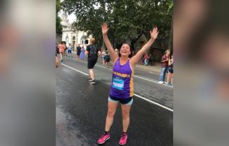 A woman in running gear standing with her hands in the air and looking exhausted but smiling