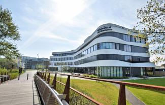 A modern university building surrounded trees and grass, with a bridge leading to it