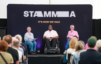 Three people sitting on a stage looking out at the audience. One of the people is in a wheelchair.