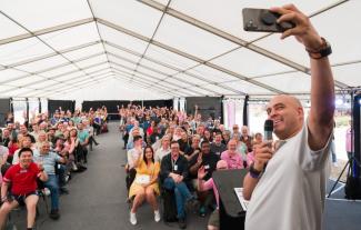 A man on a stage holding a microphone and taking a selfie with the audience