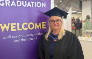 A woman in university gown and mortar board standing in front of a sign saying 'Welcome'