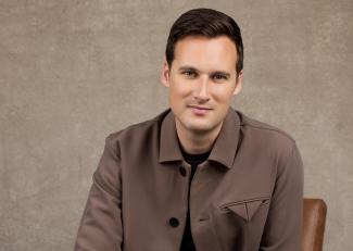 A man smiling, sitting in front of a brown background