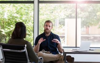 A man sitting beside a desk, with a clipboard on his lap, talking to a woman sitting opposite him.