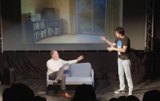 Two male actors on a stage, one sitting on a sofa and the other standing