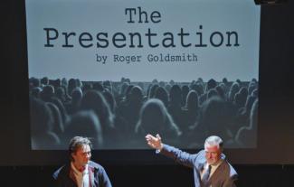 Two actors on a stage, with a backdrop saying 'The Presentation'