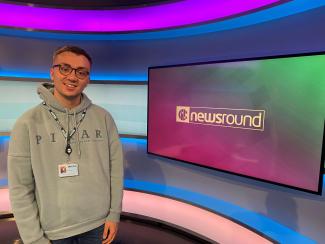 A young man in a TV studio, smiling, in front of a TV screen with the 'Newsround' logo on it