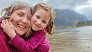A young girl embraces her mother as they both smile for the camera beside a lake