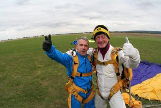 Two men in parachuting gear looking at the camera and smiling with arms around each other and their thumbs up