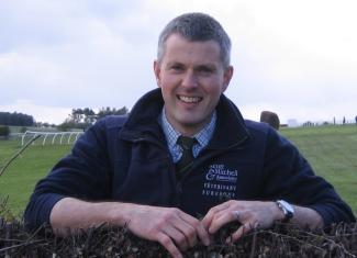 A man standing beside a hedge on a racecourse, smiling