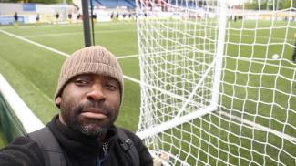 A man standing behind the goal net on a football pitch, looking at the camera