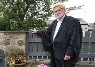 A man in a barrister's outfit leaning on a fence and smiling